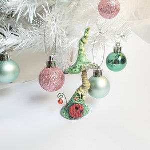 Puede incluir: Un adorno de Navidad de color verde y marrón con forma de hongo, con una puerta roja y una pequeña baya roja en un tallo. El adorno está colgado de un árbol de Navidad artificial blanco con otros adornos.