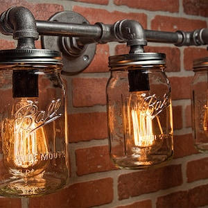 May include: Industrial-style wall sconce featuring three clear glass mason jars with Edison-style light bulbs. The jars are attached to a metal pipe fixture against a red brick wall. The jars have the word "Ball" embossed on them.