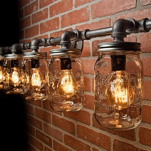Puede incluir: Una lámpara rústica hecha de tarros de cristal Mason reutilizados y tubería industrial. La lámpara tiene cinco tarros con bombillas Edison, montados en una tubería de metal contra una pared de ladrillo.
