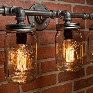 May include: Industrial-style wall sconce featuring three clear glass mason jar light fixtures. The jars are suspended from a metal pipe frame against a brick wall. Each jar contains a glowing Edison-style light bulb.