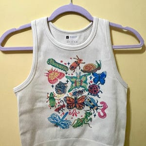 May include: A white ribbed tank top featuring a colourful graphic print of various insects, including butterflies, a snail, a ladybug, a dragonfly, and a praying mantis, arranged in a cluster with small star accents across the centre of the chest.