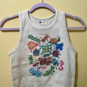 May include: Cream ribbed tank top with a vibrant insect design. The print showcases a variety of insects, such as butterflies, beetles, and a dragonfly. The top is displayed on a purple clothes hanger.
