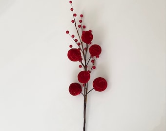 May include: A decorative floral stem with red velvet ball ornaments and small red berry accents. The stem is dark brown, and the berries and ball ornaments are a vibrant red. The arrangement is set against a white background.