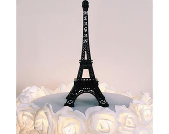 May include: A black Eiffel Tower cake topper with the name "Meagan" written on it. The topper is sitting on a white cake decorated with white roses.