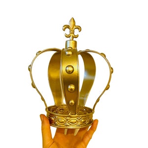 May include: A gold metal crown with a fleur-de-lis design on top. The crown has a round base and is decorated with small, round accents.