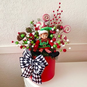 K&ouml;nnte beinhalten: Festliches Weihnachtsarrangement in einem roten Beh&auml;lter mit einer Elfenpuppe. Das Arrangement umfasst rote, gr&uuml;ne und wei&szlig;e Dekorationen, darunter Lutscher, Beeren und eine schwarz-wei&szlig; karierte Schleife. Der Elf tr&auml;gt einen gr&uuml;nen Hut und ein rot-gr&uuml;nes Outfit.