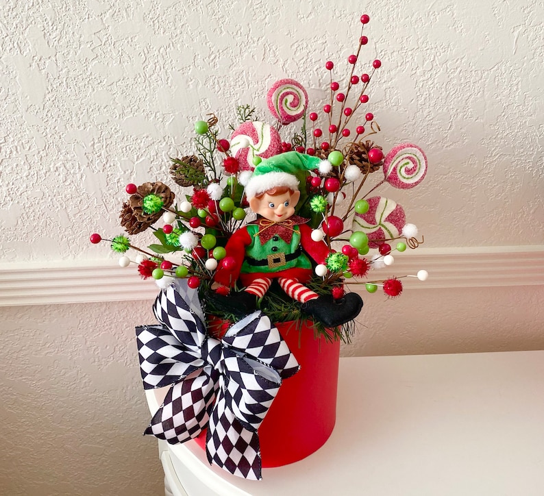 K&ouml;nnte beinhalten: Festliches Weihnachtsarrangement in einem roten Beh&auml;lter mit einer Elfenpuppe. Das Arrangement umfasst rote, gr&uuml;ne und wei&szlig;e Dekorationen, darunter Lutscher, Beeren und eine schwarz-wei&szlig; karierte Schleife. Der Elf tr&auml;gt einen gr&uuml;nen Hut und ein rot-gr&uuml;nes Outfit.