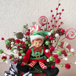 K&ouml;nnte beinhalten: Weihnachtliche Elfendekoration in einem roten Topf, umgeben von roten Beeren, gr&uuml;nen und wei&szlig;en Kugeln und zuckerstangenf&ouml;rmigen Dekorationen. Der Elf tr&auml;gt einen gr&uuml;nen Hut und ein rot-gr&uuml;nes Outfit. Eine schwarz-wei&szlig; karierte Schleife schm&uuml;ckt den Topf.