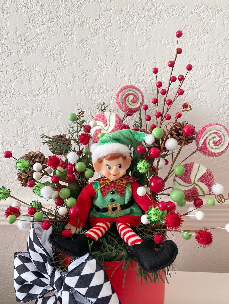 K&ouml;nnte beinhalten: Weihnachtliche Elfendekoration in einem roten Topf, umgeben von roten Beeren, gr&uuml;nen und wei&szlig;en Kugeln und zuckerstangenf&ouml;rmigen Dekorationen. Der Elf tr&auml;gt einen gr&uuml;nen Hut und ein rot-gr&uuml;nes Outfit. Eine schwarz-wei&szlig; karierte Schleife schm&uuml;ckt den Topf.
