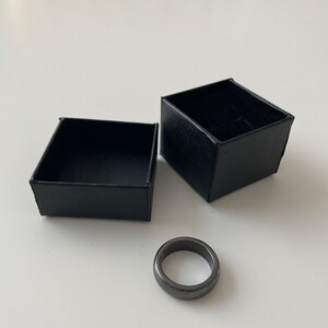 May include: A black ring made of hematite in a black ring box. The ring is smooth and has a polished finish.