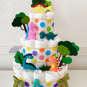 May include: A three-tiered diaper cake decorated with colorful polka dots and dinosaur figurines. The cake is topped with a green felt tree and a yellow dinosaur figurine.