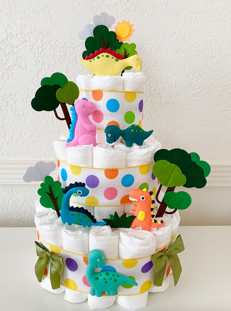 May include: A three-tiered diaper cake decorated with colorful polka dots and dinosaur figurines. The cake is topped with a green felt tree and a yellow dinosaur figurine.