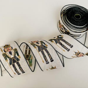 May include: White ribbon with a black trim featuring a repeating pattern of nutcracker soldiers in black, gold, and green. A roll of the same ribbon is visible in the background. This decorative ribbon is perfect for holiday crafts.