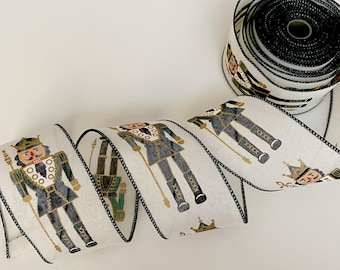 May include: White ribbon with a black trim featuring a repeating pattern of nutcracker soldiers in black, gold, and green. A roll of the same ribbon is visible in the background. This decorative ribbon is perfect for holiday crafts.
