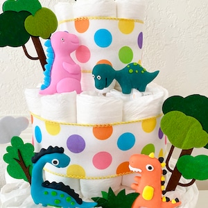 May include: A multi-tiered diaper cake decorated with colorful polka dot ribbon and dinosaur figurines. The cake features a green, blue, pink, and yellow dinosaur, as well as green felt trees.