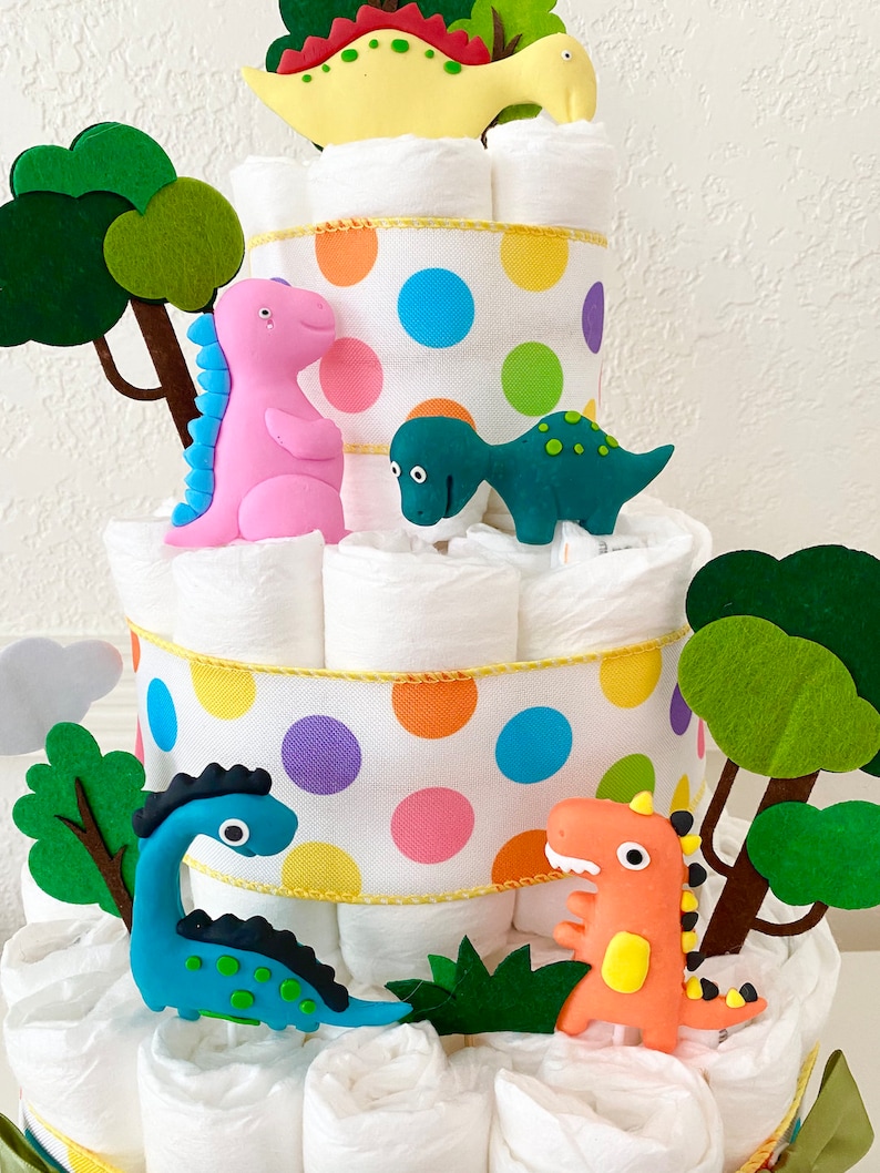 May include: A multi-tiered diaper cake decorated with colorful polka dot ribbon and dinosaur figurines. The cake features a green, blue, pink, and yellow dinosaur, as well as green felt trees.