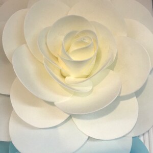 May include: A white paper flower with multiple layers of petals, creating a 3D effect. The flower is made of white paper circles, layered and curled to form a rose shape.