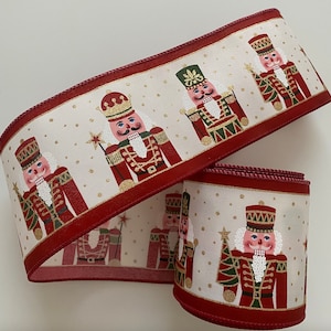 May include: Cream-colored ribbon featuring a repeating pattern of nutcracker soldiers in red, green, and gold. The ribbon has a red border and is dotted with gold specks, perfect for holiday crafts and decorations.