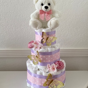May include: A three-tier diaper cake decorated with pink and purple ribbon, white polka dots, and gold butterflies. A white teddy bear with a pink bow sits on top of the cake.