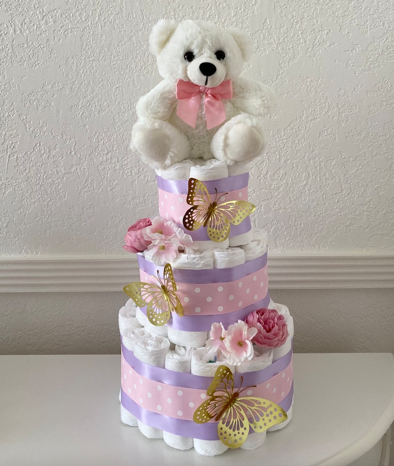 May include: A three-tier diaper cake decorated with pink and purple ribbon, white polka dots, and gold butterflies. A white teddy bear with a pink bow sits on top of the cake.