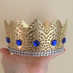 May include: Gold crown with a delicate lace design and blue gem accents. The crown has a silver trim around the base.