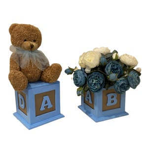 May include: Two blue wooden blocks with brown letters spelling "A" and "B". One block has a brown teddy bear sitting on top of it. The other block has a bouquet of white and blue artificial flowers.