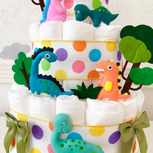 May include: A three-tiered diaper cake decorated with colorful polka dots and green felt trees. The cake is adorned with five dinosaur figurines, including a pink, green, blue, and two orange dinosaurs.
