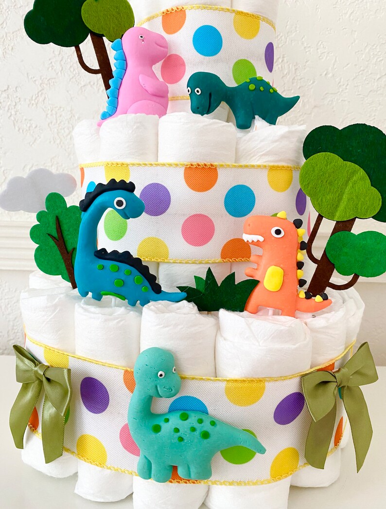 May include: A three-tiered diaper cake decorated with colorful polka dots and green felt trees. The cake is adorned with five dinosaur figurines, including a pink, green, blue, and two orange dinosaurs.