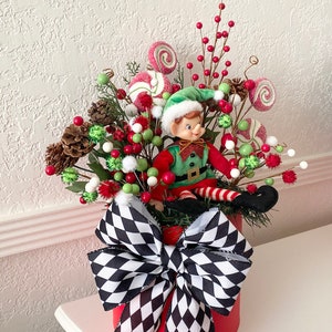 K&ouml;nnte beinhalten: Festliche Weihnachtsdekoration in einem roten Beh&auml;lter, verziert mit einer gro&szlig;en schwarz-wei&szlig; karierten Schleife. Sie enth&auml;lt eine Elfenfigur, rote und gr&uuml;ne Beeren, Tannenzapfen und wirbelnde S&uuml;&szlig;igkeiten-Dekorationen.