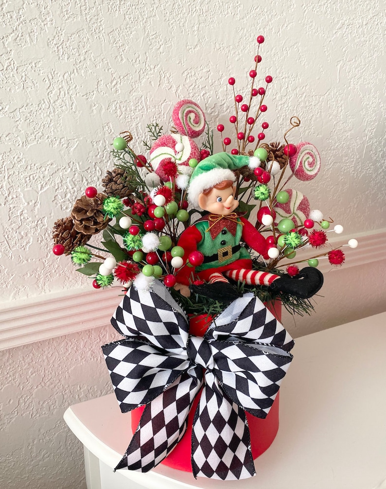 K&ouml;nnte beinhalten: Festliche Weihnachtsdekoration in einem roten Beh&auml;lter, verziert mit einer gro&szlig;en schwarz-wei&szlig; karierten Schleife. Sie enth&auml;lt eine Elfenfigur, rote und gr&uuml;ne Beeren, Tannenzapfen und wirbelnde S&uuml;&szlig;igkeiten-Dekorationen.