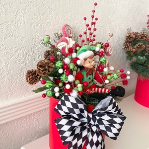 K&ouml;nnte beinhalten: Festliche Weihnachtsdekoration in einem roten Zylinder. Sie enth&auml;lt eine Elfenfigur, Tannenzapfen, rote Beeren, gr&uuml;ne und wei&szlig;e Ornamente und Lutscher. Eine gro&szlig;e schwarz-wei&szlig; karierte Schleife schm&uuml;ckt die Vorderseite und sorgt f&uuml;r eine fr&ouml;hliche Atmosph&auml;re.