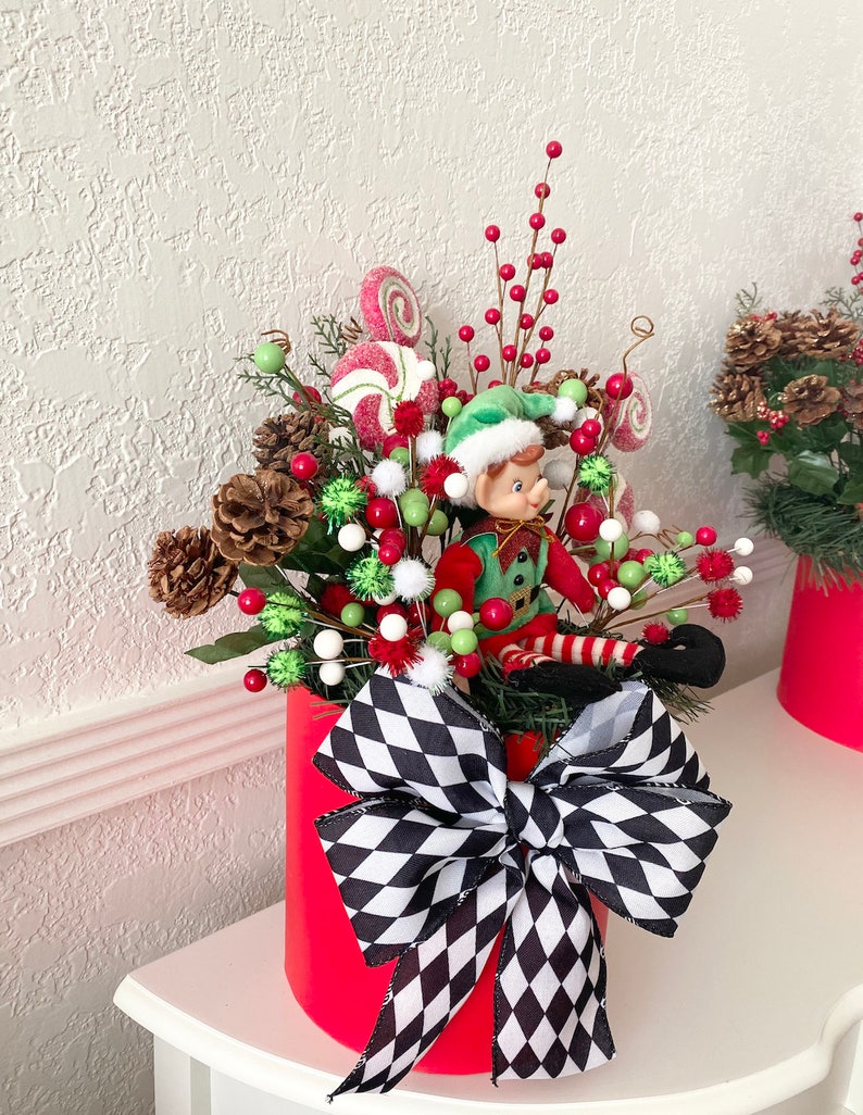 K&ouml;nnte beinhalten: Festliche Weihnachtsdekoration in einem roten Zylinder. Sie enth&auml;lt eine Elfenfigur, Tannenzapfen, rote Beeren, gr&uuml;ne und wei&szlig;e Ornamente und Lutscher. Eine gro&szlig;e schwarz-wei&szlig; karierte Schleife schm&uuml;ckt die Vorderseite und sorgt f&uuml;r eine fr&ouml;hliche Atmosph&auml;re.