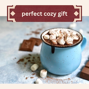 Może przedstawiać: Jasnoniebieski, nakrapiany kubek ceramiczny wypełniony gorącą czekoladą z białymi piankami marshmallow, z ciemnoczerwonym banerem u g&oacute;ry z białym napisem perfect cozy gift, w towarzystwie rozrzuconych kawałk&oacute;w czekolady i pianek marshmallow.