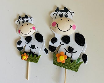 May include: Two black and white cow cake toppers with green grass and orange flower accents. The larger cow is standing with its head tilted to the side, while the smaller cow is sitting with its legs crossed.