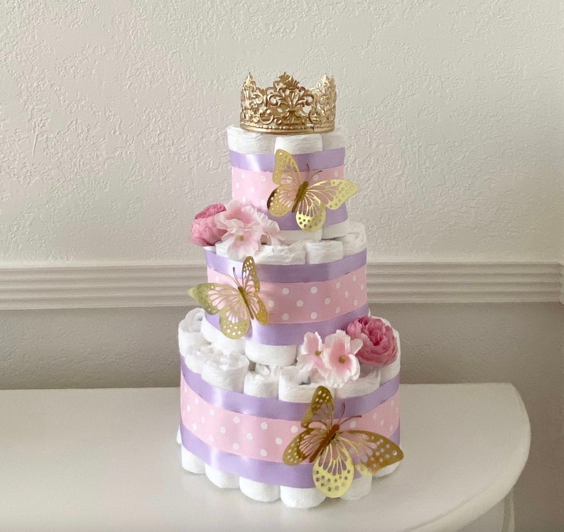 May include: A three-tiered diaper cake decorated with pink and purple ribbon, white polka dots, and gold butterflies. The top tier is topped with a gold crown.