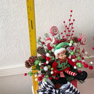 K&ouml;nnte beinhalten: Weihnachtself-Dekoration mit einem rot-gr&uuml;n gekleideten Elf, umgeben von S&uuml;&szlig;igkeiten-Ornamenten, Tannenzapfen und roten Beeren. Die Anordnung befindet sich in einem roten Beh&auml;lter mit einer schwarz-wei&szlig; karierten Schleife. Die H&ouml;he betr&auml;gt ca. 56 cm.