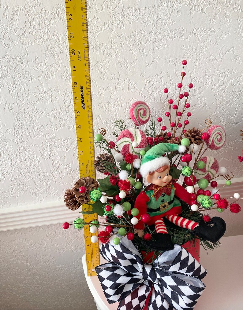 K&ouml;nnte beinhalten: Weihnachtself-Dekoration mit einem rot-gr&uuml;n gekleideten Elf, umgeben von S&uuml;&szlig;igkeiten-Ornamenten, Tannenzapfen und roten Beeren. Die Anordnung befindet sich in einem roten Beh&auml;lter mit einer schwarz-wei&szlig; karierten Schleife. Die H&ouml;he betr&auml;gt ca. 56 cm.