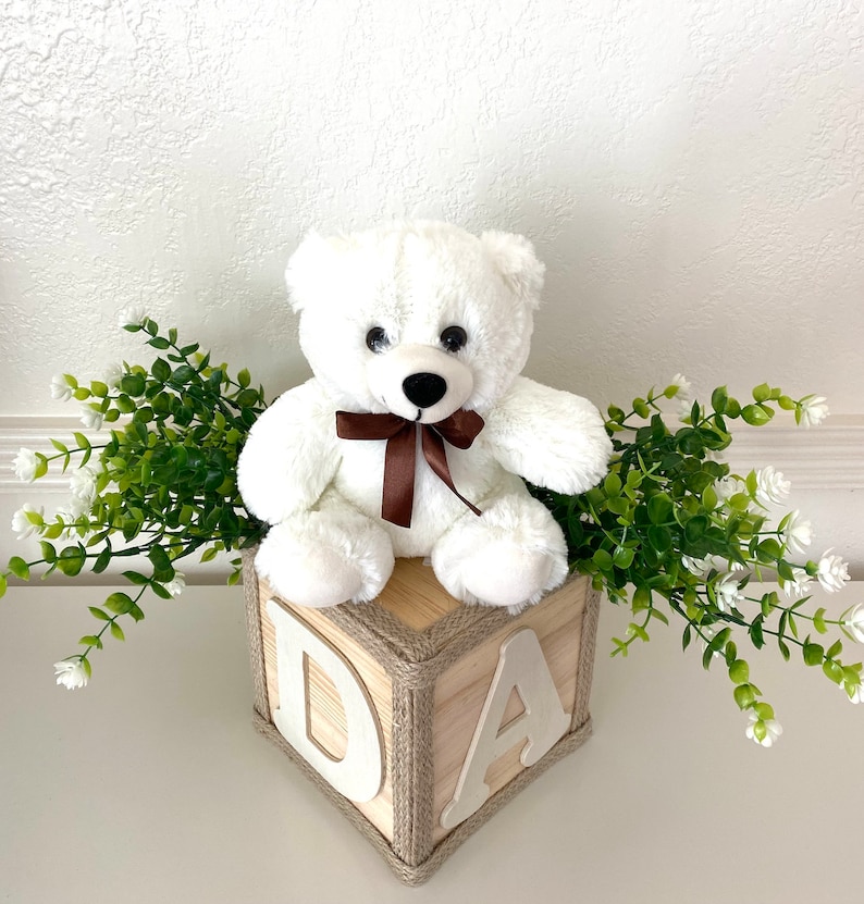 May include: White teddy bear sitting on a wooden block with the letters "DA" on it. The block is wrapped in twine and the bear is wearing a brown ribbon. There are green plants with white flowers on either side of the block.
