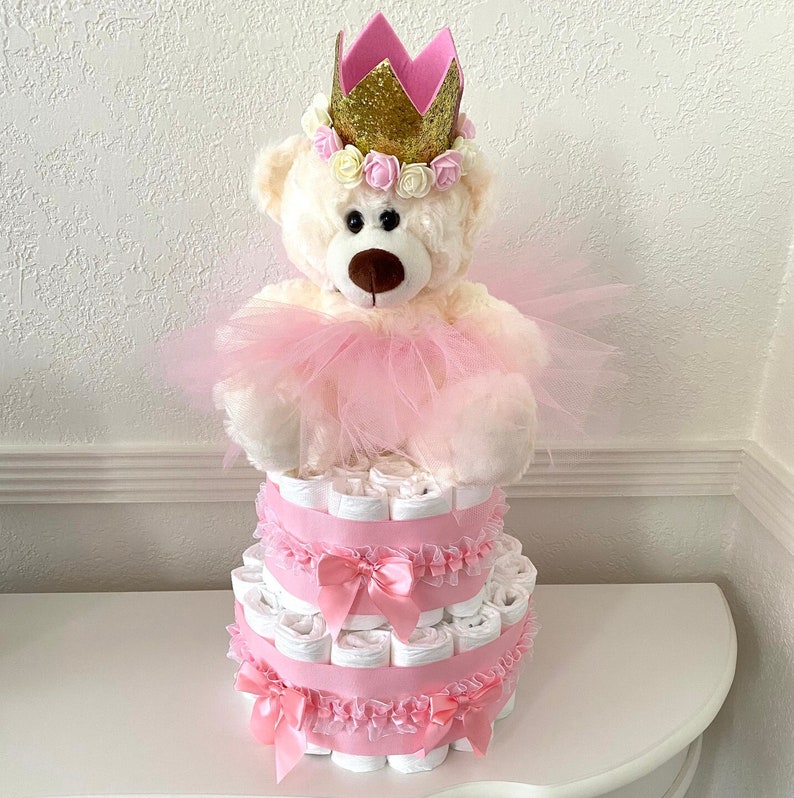 May include: A two-tier diaper cake decorated with pink tulle, ribbons, and bows. A white teddy bear wearing a pink tutu and a gold crown with pink flowers sits on top of the cake.