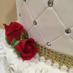 May include: A white wedding cake decorated with a quilted pattern and rhinestones. The bottom tier has a gold and white ribbon with a row of pearls. Two red roses are placed on the side of the cake.