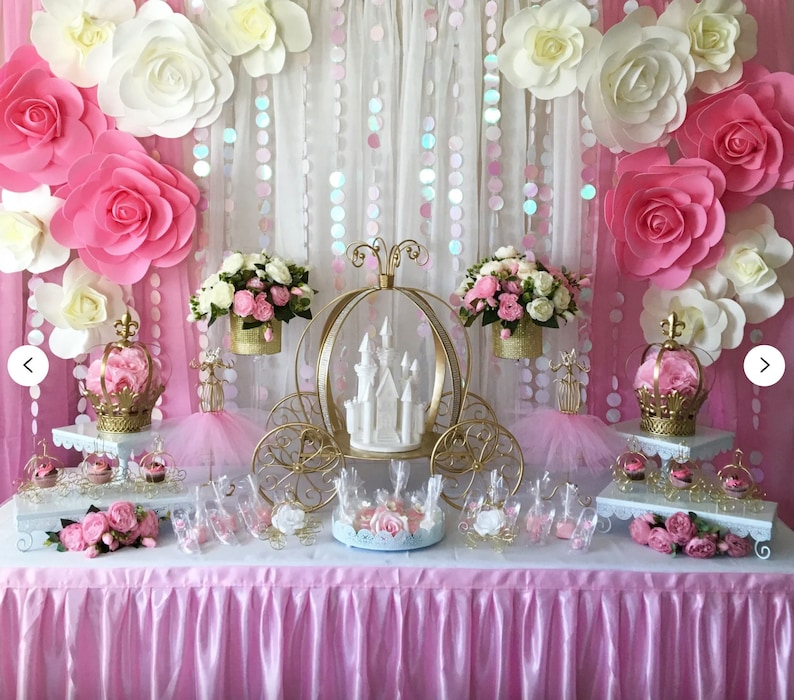 May include: A pink and white princess themed party dessert table with a gold carriage centrepiece, white cake, pink and white flowers, and cupcakes.