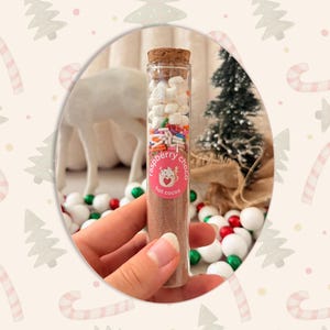 Może przedstawiać: Przezroczysta szklana prob&oacute;wka wypełniona brązowym proszkiem do gorącej czekolady, zwieńczona białymi mini piankami marshmallow i kolorową posypką. Okrągła r&oacute;żowa etykieta z przodu zawiera ilustrację kota oraz napis raspberry choco hot cocoa.