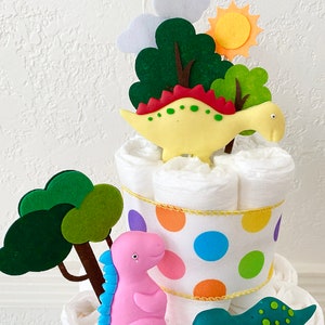 May include: A diaper cake decorated with felt trees, a sun, and three dinosaur figurines. The dinosaurs are yellow, pink, and green. The cake is wrapped in a white ribbon with colorful polka dots.