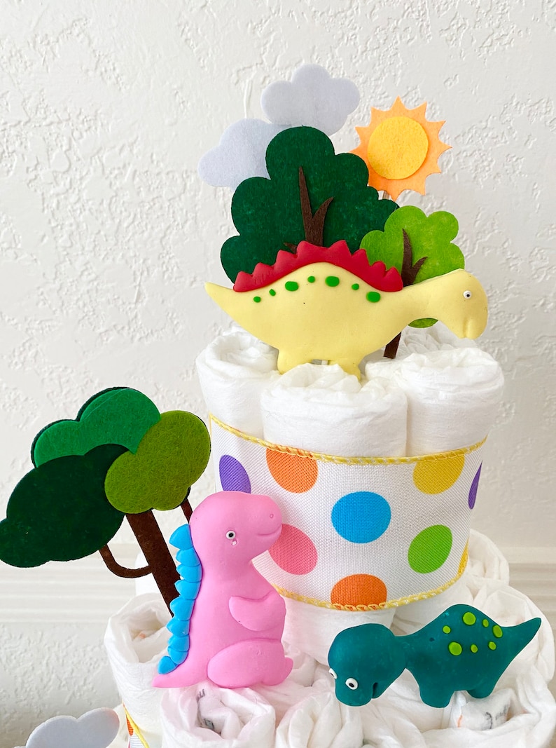 May include: A diaper cake decorated with felt trees, a sun, and three dinosaur figurines. The dinosaurs are yellow, pink, and green. The cake is wrapped in a white ribbon with colorful polka dots.