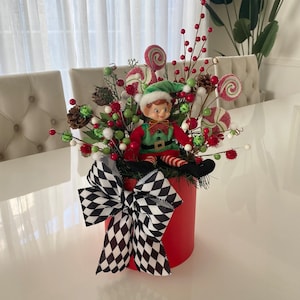 K&ouml;nnte beinhalten: Ein festliches Weihnachtsgesteck in einem roten, zylindrischen Beh&auml;lter. Es zeigt eine Elfenfigur, rote und gr&uuml;ne Beeren, Tannenzapfen und gewirbelte Lutscher. Eine gro&szlig;e schwarz-wei&szlig;e karierte Schleife schm&uuml;ckt die Vorderseite und sorgt f&uuml;r eine fr&ouml;hliche Weihnachtsdekoration.