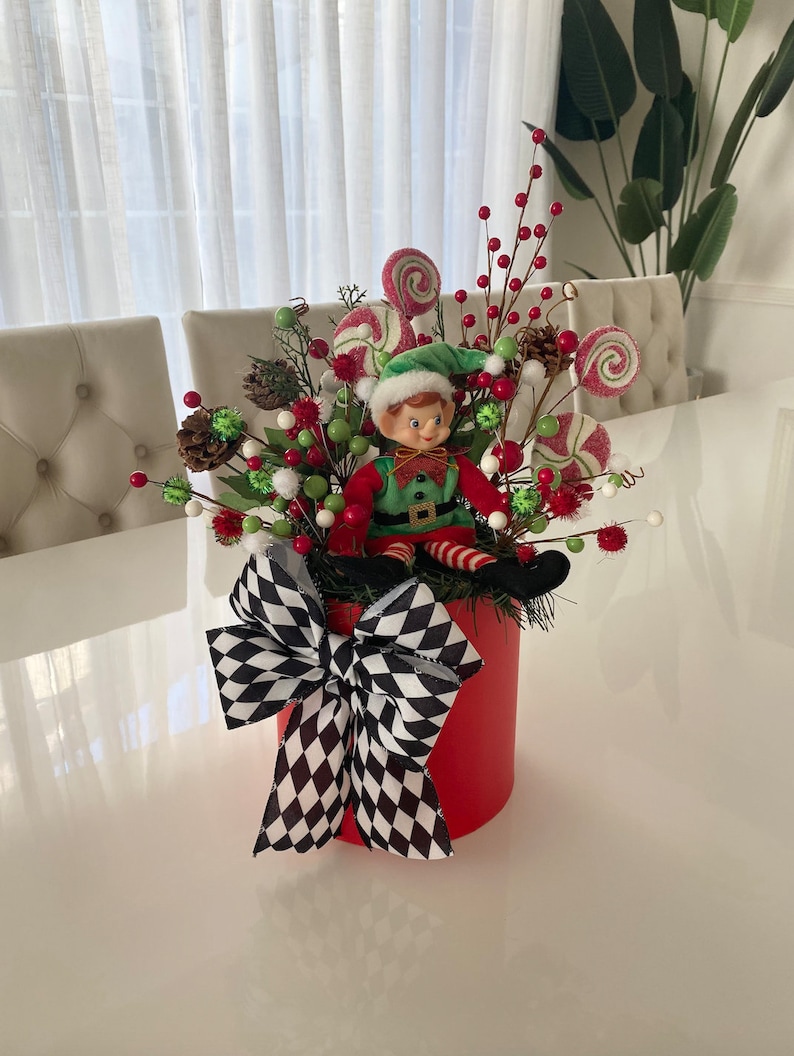 K&ouml;nnte beinhalten: Ein festliches Weihnachtsgesteck in einem roten, zylindrischen Beh&auml;lter. Es zeigt eine Elfenfigur, rote und gr&uuml;ne Beeren, Tannenzapfen und gewirbelte Lutscher. Eine gro&szlig;e schwarz-wei&szlig;e karierte Schleife schm&uuml;ckt die Vorderseite und sorgt f&uuml;r eine fr&ouml;hliche Weihnachtsdekoration.