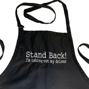 May include: Black apron with a large pocket and adjustable straps. The apron has white text that reads "Stand Back! I'm taking out my driver."