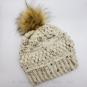May include: A cream-coloured crocheted beanie with a brown faux fur pom pom. The beanie has a textured stitch pattern and a ribbed brim.
