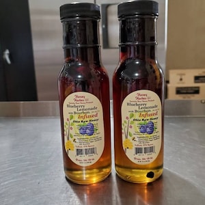 May include: Two glass bottles of honey infused blueberry lemonade with bourbon. The bottles are labeled "Honey Hostess LLC Blueberry Lemonade with Bourbon Infused Ohio Raw Honey" and have a blue and white label with a blueberry graphic. The bottles are on a stainless steel surface.