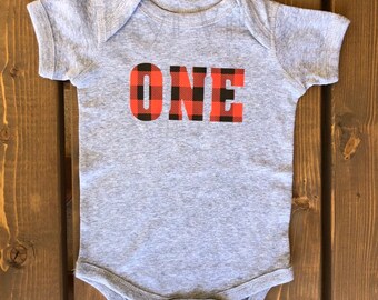 Op de afbeelding: Een grijze baby romper met het woord "ONE" in zwarte letters met een rood-zwart buffalo plaid patroon.